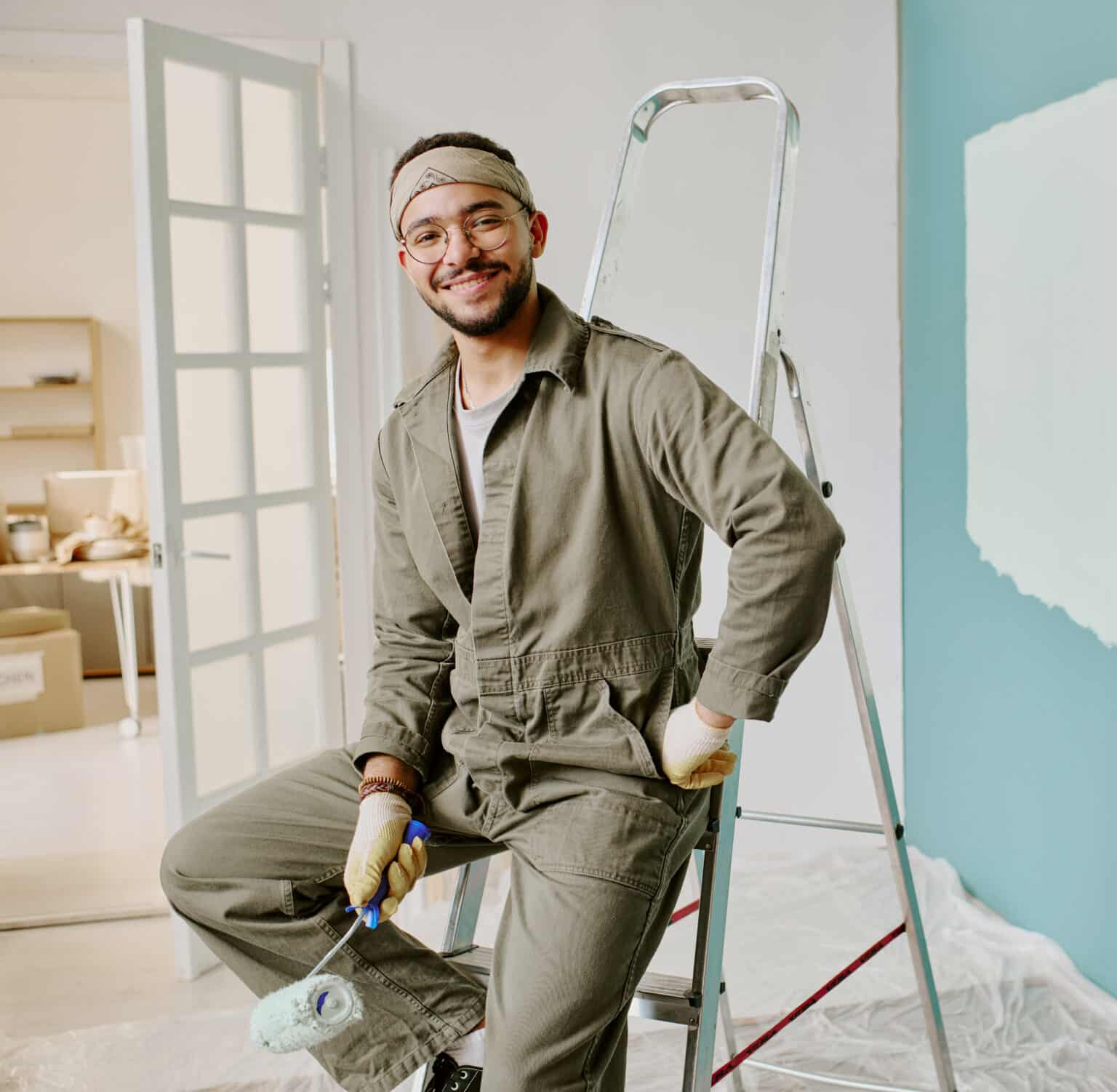 Smiling interior house painter Edmonton in coveralls with roller on ladder beside a teal wall being repainted in Highlands.
